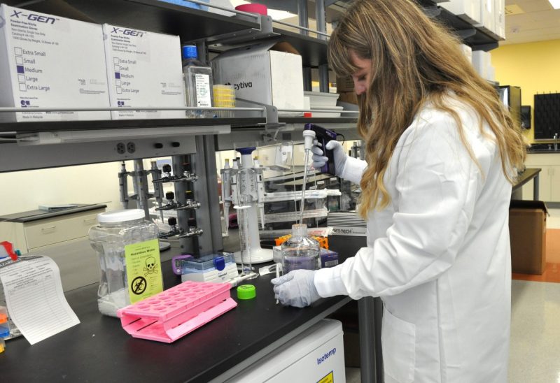 Female student, with long blonde hair, wearing a white lab coat, works in a lab